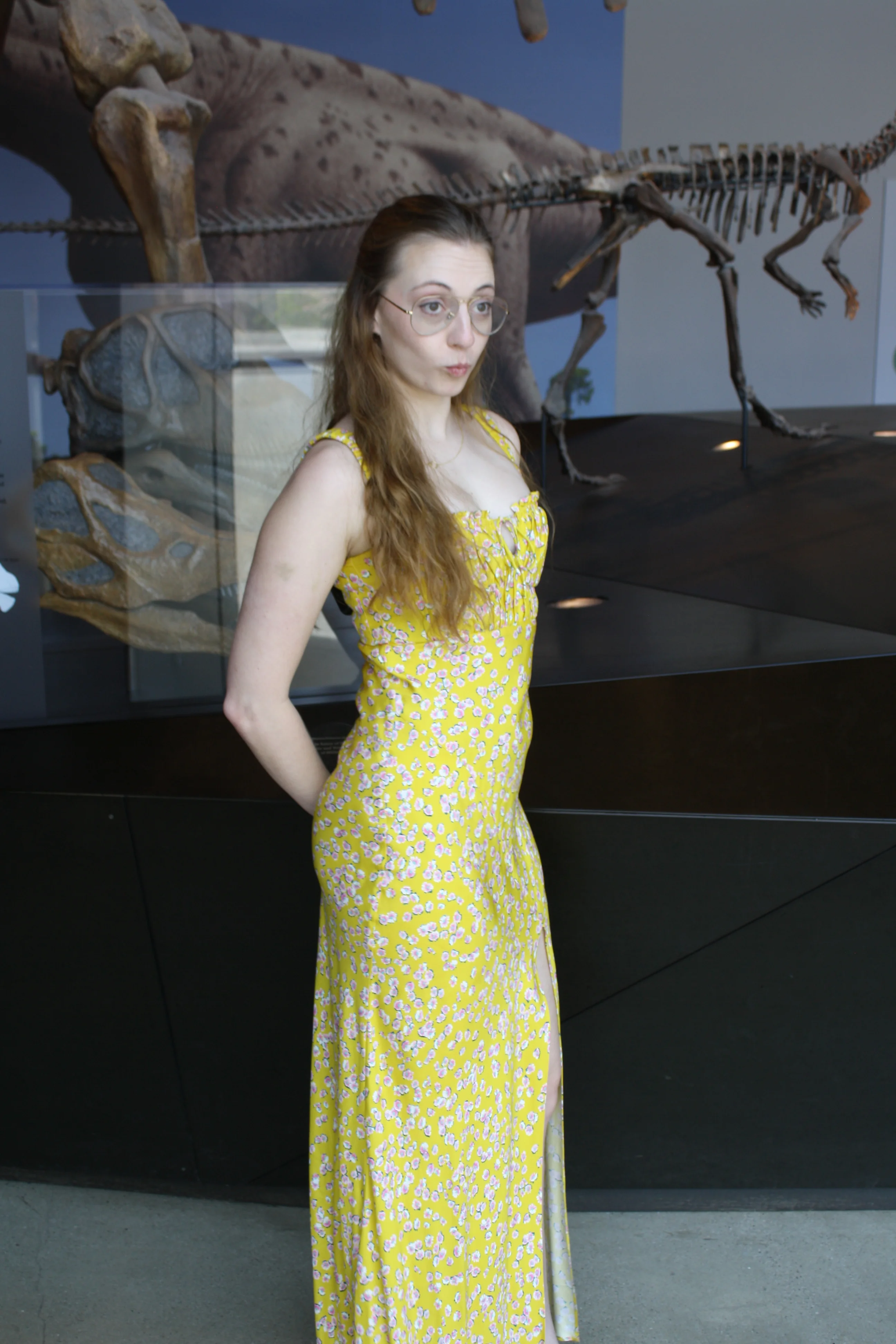 Jordie Morris standing in a yellow floral dress beside a museum dinosaur exhibit.